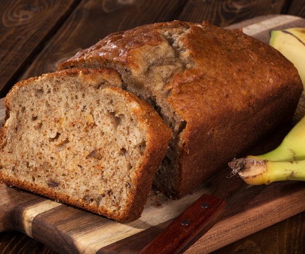 Closeup of a slice of banana nut bread on a wooden cutting board with two bananas on the side