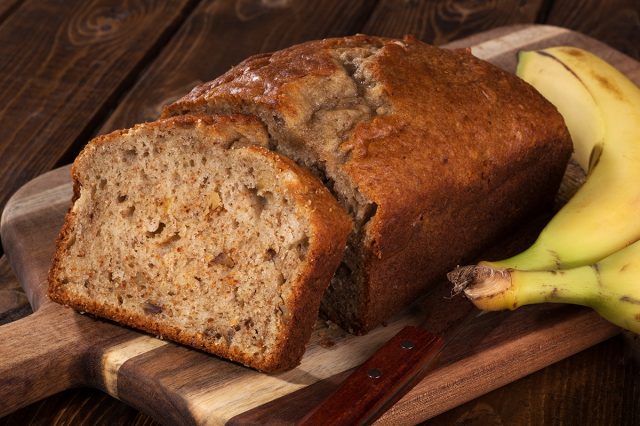 Closeup of a slice of banana nut bread on a wooden cutting board with two bananas on the side