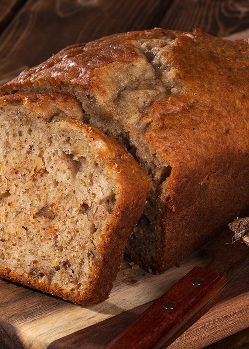 Closeup of a slice of banana nut bread on a wooden cutting board with two bananas on the side