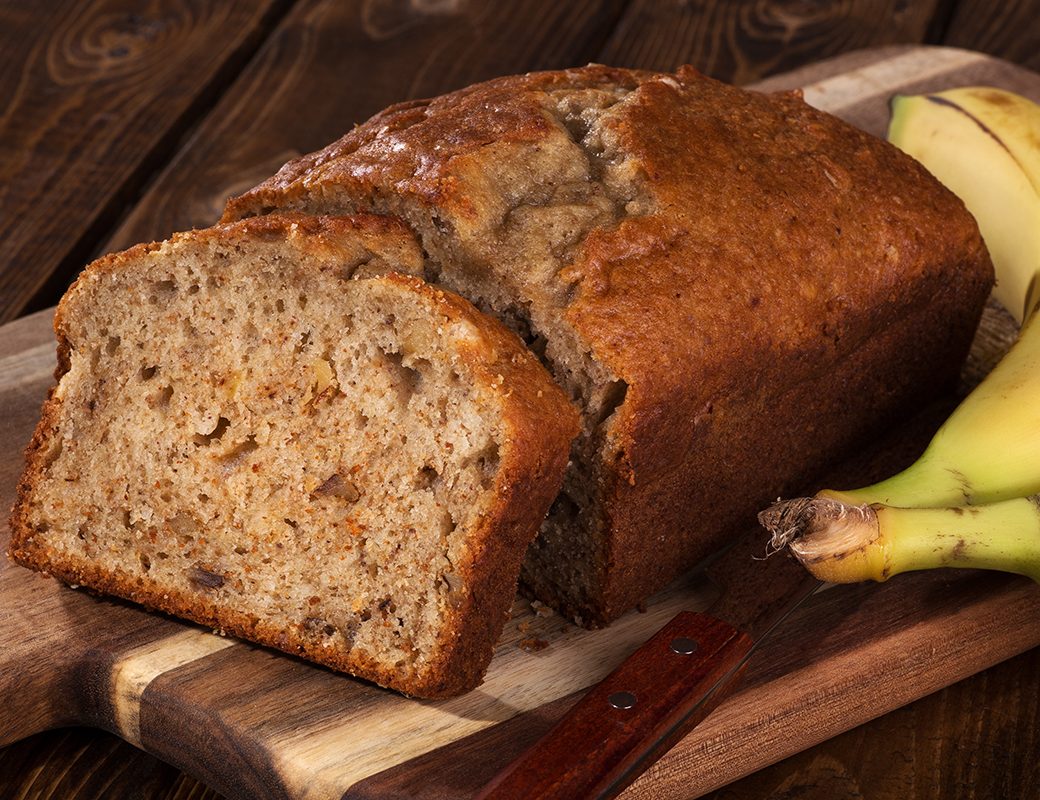 Closeup of a slice of banana nut bread on a wooden cutting board with two bananas on the side