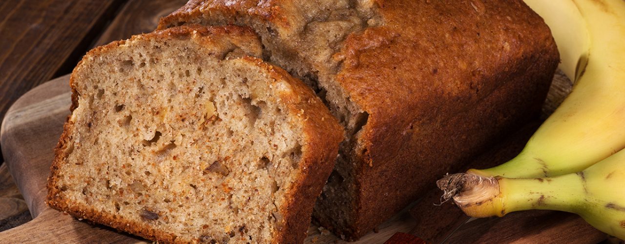 Closeup of a slice of banana nut bread on a wooden cutting board with two bananas on the side
