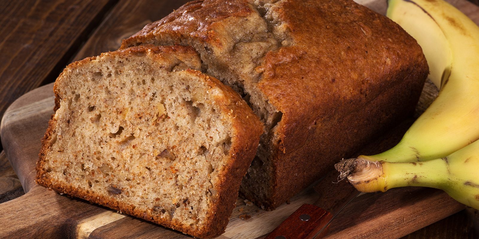 Closeup of a slice of banana nut bread on a wooden cutting board with two bananas on the side