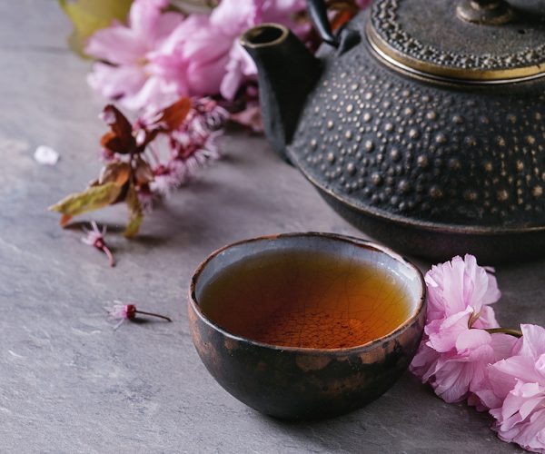 Black iron teapot and traditional ceramic cup of tea with blossom pink flowers cherry branch over gray texture background. Asian style.
